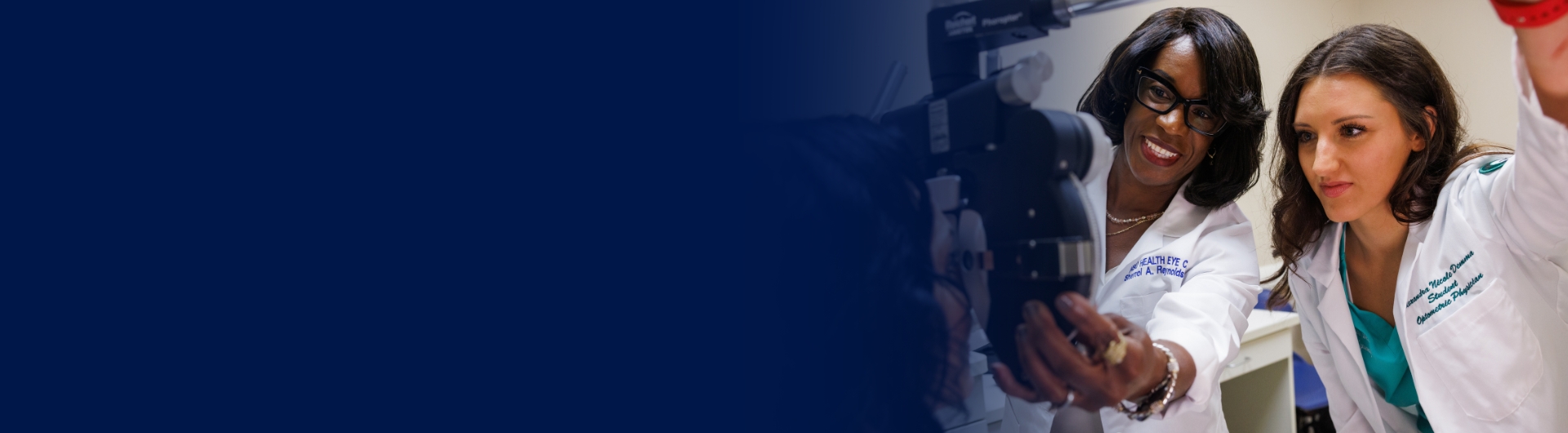 An eye health exam room with two females, including one optometry student, looking an eye care equipment.