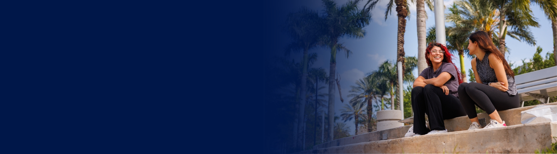 Two female students talking on outdoor steps on a campus with palm trees in the background.