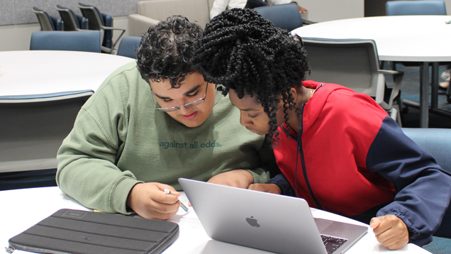 Two students at a table leaning over paper with laptop in front of them