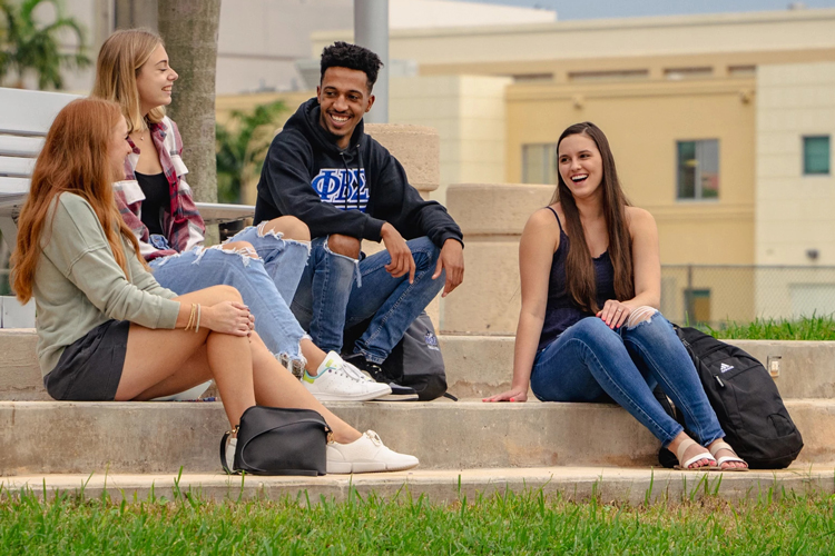 Four students sitting on the steps outside a building, smiling and talking