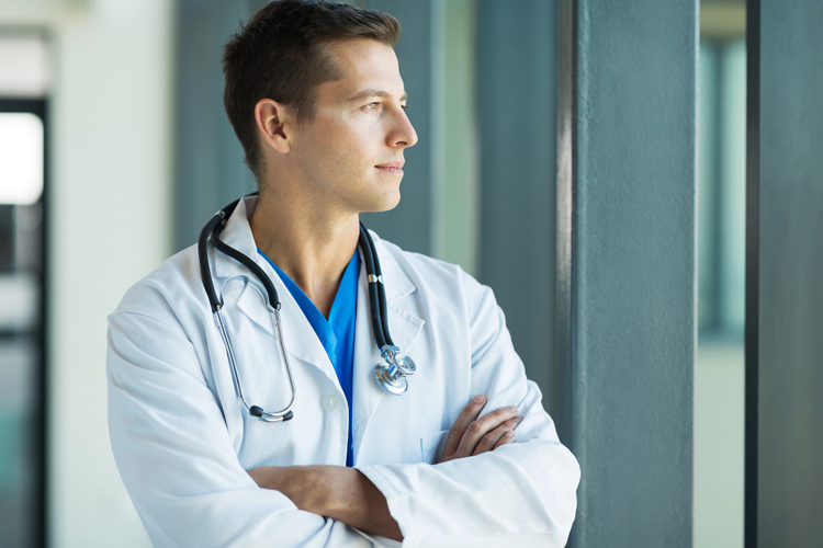 Young doctor wearing white coat and stethoscope looking out window.