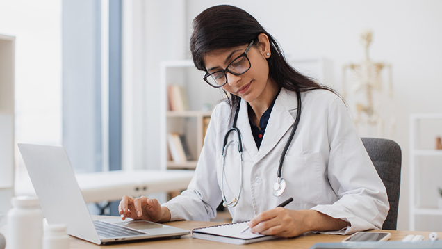 Doctor writing in a notebook while using a laptop