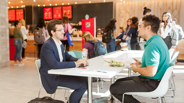 Two students in the foreground sitting at a table in the food court.